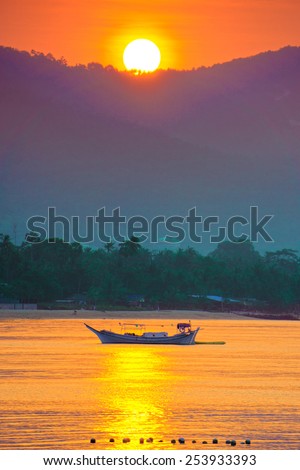 The sun sets behind the mountain, lighting up the last rays of a fishing boat, which is anchored in a tropical bay