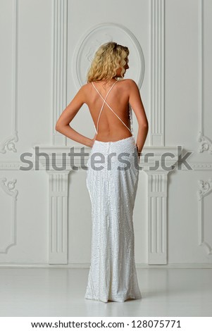 Beautiful blonde woman in sexy white long dress with naked back. Shooting in white interior studio.