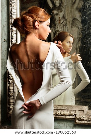 Portrait of the beautiful woman in white dress with naked back. She is standing at the mirror. Studio with interior of old palace. Not necessary property release.