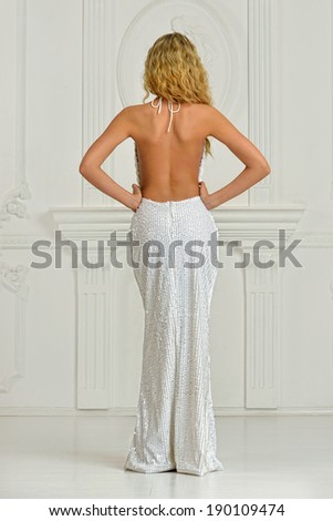 Beautiful blonde woman in sexy white long dress with naked back. Shooted in white interior studio.