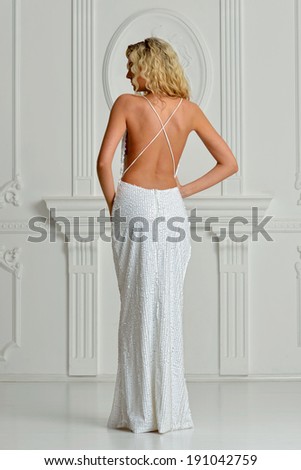 Beautiful blonde woman in sexy white long dress with naked back. Shooted in white interior studio.