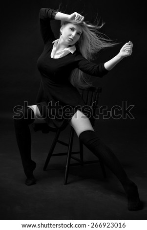 Beautiful woman in a black dress and stockings posing on the chair in studio with dark background .