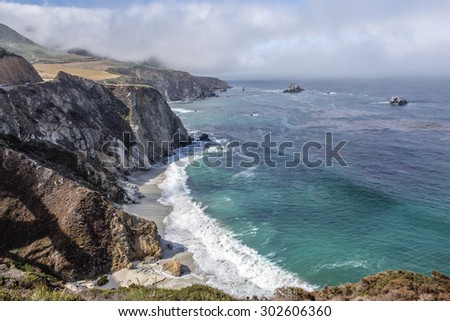 Waves splashing on huge rocks, off shore, along a rocky coastline,fog & cloud covered mountain tops, bridge, traveling the Big Sur Highway (Highway 1), on the California Central Coast.