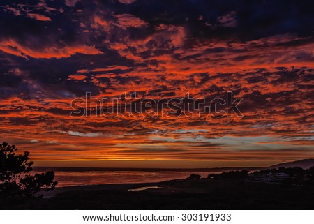 Burning red sun, thunderheads, pending storm, painted sky, and reflective sea at sunset, on (Big Sur Coast) Moonstone Beach, with people in the distance. California Central Coast, near Cambria CA.