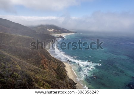 Waves splashing on huge rocks, off shore, along a rocky coastline, fog & cloud covered mountain tops, bixby bridge, traveling the Big Sur Highway (Highway 1), on the California Central Coast.