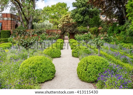 A hidden gate is opening up to a beautiful English style garden with rounded hedges, beautiful flowers, & symmetric type design, with sand & brick sidewalks and footpaths to enjoy the scenic view.