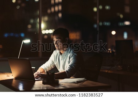 Young Asian businessman working on a laptop while sitting at his office desk at night with city lights in the background