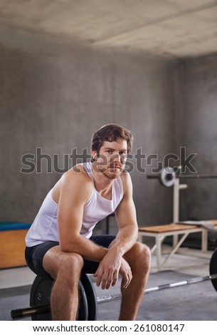 Serious young man looking at the camera while sitting on a weight in his private gym