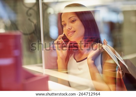 A young woman looking at a handbag through a shop window while talking on the phone