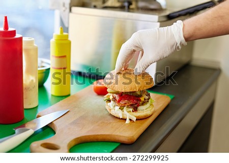 Cropped shot of a white latex gloved hand carefully putting the top bun down on a big delicious burger that\'s been freshly prepared in a kitchen