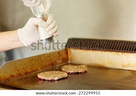 Cropped shot of gloved cook\'s hands grinding salt onto two hamburger patties to season them while they are frying on the grill