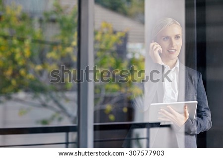 A young businesswoman seen talking on the phone through her office window, while holding touchpad
