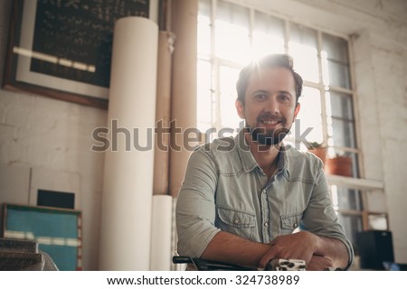Portrait of a handsome young male designer looking positively at the camera with gentle sun flare coming in through the window of his studio