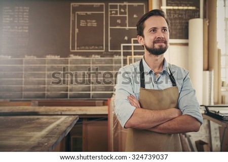 Cratsman looking away in a thoughtful manner with his arms folded while standing in his carpentry workshop and smiling to himself