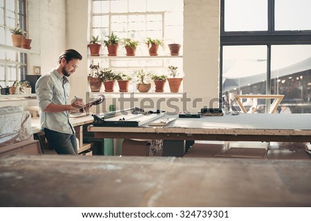 Young male business owner standing in his studio workshop checking figures and stock on his clipboard on a sunny afternoon