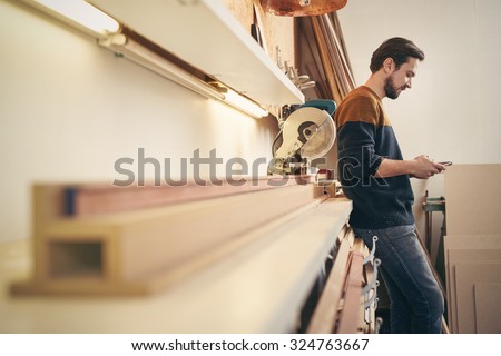 Craftsman leaning against a work area in his workshop using his cell phone to send a message