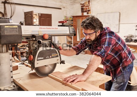 Skilled carpenter in his woodwork workshop, cutting a piece of wood with a radial arm circular saw, and wearing safety goggles