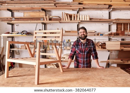 Portrait of a smiling craftsman with a rugged beard, resting on his workbench with a wooden chair frame on it, looking at the camera, with shelves of wooden planks and pieces behind him