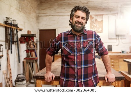 Portrait of a smiling craftsman with a rugged beard wearing a checked shirt, standing in his woodwork studio with a wooden workbench and tools on the wall