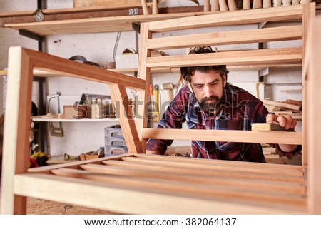 Owner of an independent furniture design and manufacturing business, working on a wooden chair frame that he has made in his woodwork studio, carefully sanding it smooth