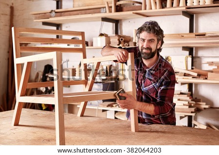 Portrait of an artisan designer, with new piece of furniture, finishing off the sanding of the chair in his studio, with shelves of wood behind him