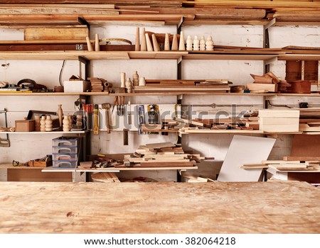 Woodwork workshop wall with many shelves holding a variety of wooden pieces and planks of wood, and some hand tools, with a wooden work bench in the foreground