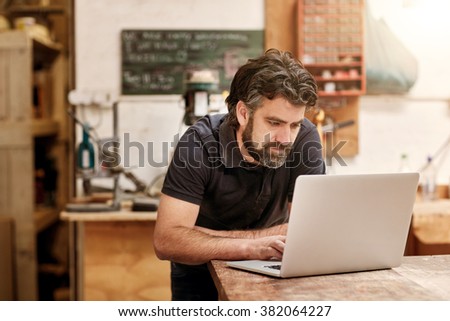 Male designer and craftsman with a rugged beard, working on his laptop at his workbench, in his studio workshop