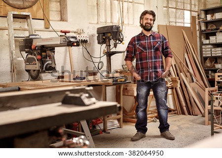 Full length portrait of a business owner who runs a carpentry studio, standing confidently with his hands in his pockets, smiling at the camera, in workshop with pieces of wood and woodwork machinery