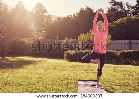 Athletic senior woman standing confidently in a balancing pose on her yoga mat, in her garden on the grass, with morning summer sunflare filtering through the trees