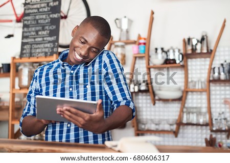 Smiling young African entrepreneur talking on a cellphone and using a digital tablet while standing at the counter of his cafe