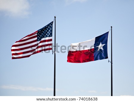 The American and Texas flags flying high together against a blue lightly cloudy sky.