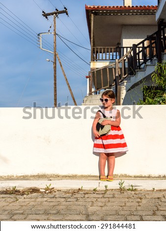 Little girl in sunglasses with a slight smile on her face dressed in a striped dress standing with a bag over her shoulder looking aside