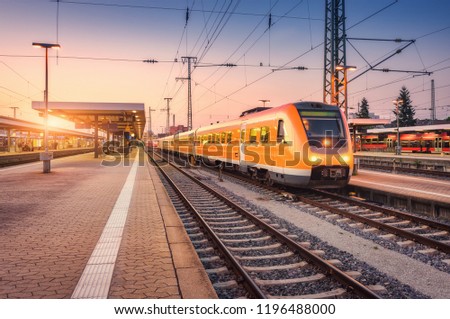 Orange high speed train on the railway station at sunset. Urban landscape with modern commuter train on the railway platform against colorful sky at dusk. Passenger vehicle on railroad in Europe