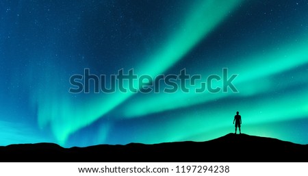 Aurora and silhouette of alone standing man on the hill. Lofoten islands, Norway. Aurora borealis and young man. Sky with stars and green polar lights. Night landscape with northern lights. Concept
