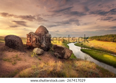 Beautiful summer sunset at the river with orange sky, clouds, rocks, yellow grass, flowers and water with reflection. Ukraine