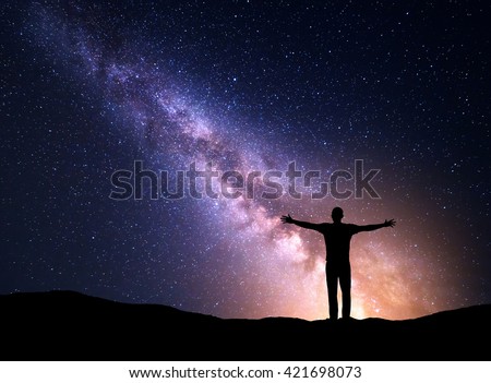 Night colorful landscape with Milky Way, yellow city lights and silhouette of a standing sporty man with raised up arms on the mountain. Universe. Travel background with purple sky full of stars