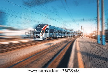 High speed train in motion at the railway station at sunset in Europe. Modern intercity train on railway platform with motion blur effect. Industrial landscape. Passenger train on railroad. Vintage