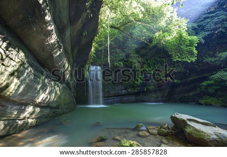 A cool refreshing waterfall and an emerald pond in a mysterious forest of lush greenery with sunlight leakage