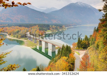 A curved bridge crossing over Lake Sylvenstein with beautiful reflections on the smooth lake water ~ Autumn scenery of Bavaria, Germany