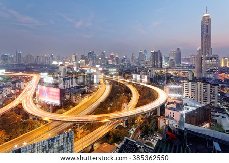 Panorama of Bangkok at dusk with skyscrapers in background and traffic trails on elevated expressways and circular interchanges ~ Bangkok at rush hour with busy traffic on intertwined highway overpass
