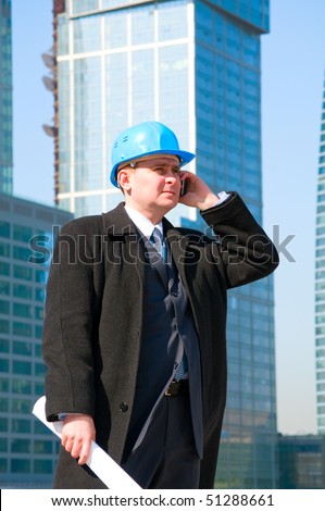 Businessman with hard hat talking on cell phone and holding drawing in his hand,on skyscrapers background