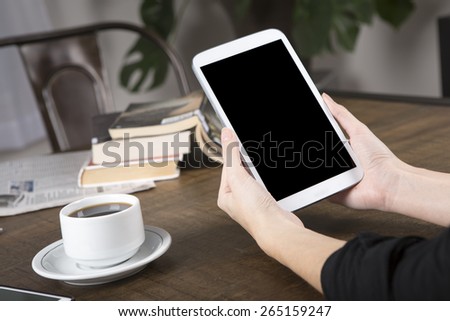 Photo of a woman holding a tablet with no name coffee cup and a stack of books in background on a wood table with shallow depth of field