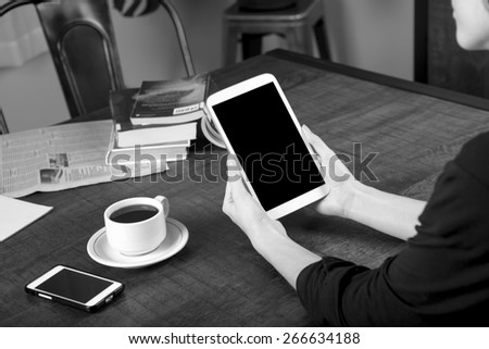 Black and white photo of a woman holding a tablet with a no name coffee cup and a stack of books in background on a wood table with shallow depth of field