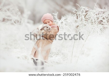 little cute smiling girl in pink hat and beige coat standing in the winter magic forest and holding a rabbit in her hands