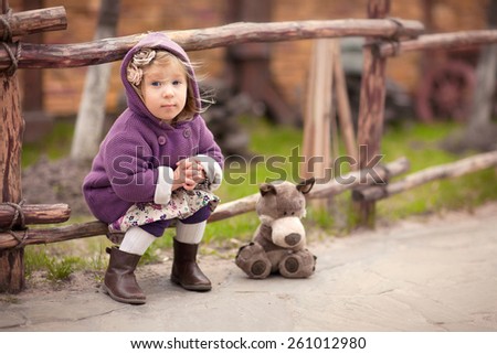 little cute girl in the purple coat with hood is sitting near the wooden fence with her toy wolf