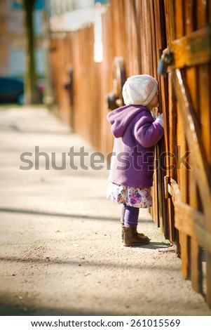 little girl in the purple coat and cream hat is looking through a hole in the wooden fence