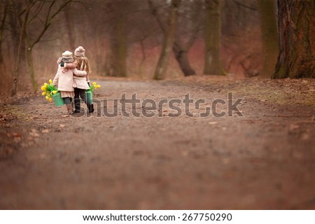 two little girls friends  two green cans with yellow flowers tulips are walking on the road hugging in the park or forest