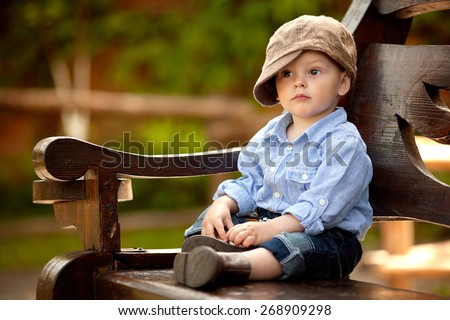 little boy in the blue shirt and brown cap is sitting on the wooden bench in the park