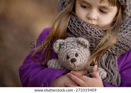 portrait of little girl in brown hat and scarf and purple coat holding teddy bear in her hands in the park