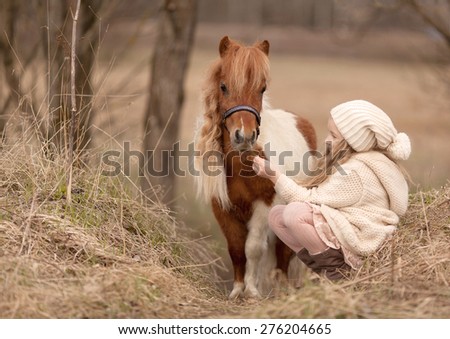 little blond girl with long hair in cream hat and coat is sitting near the little horse and feed her